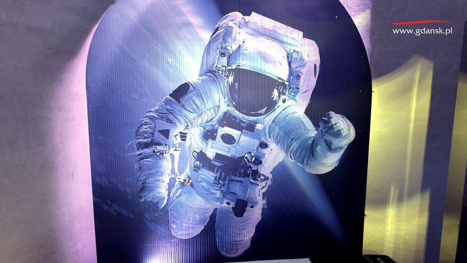 Na zdjęciu widoczna jest realistyczna grafika astronauty unoszącego się w przestrzeni kosmicznej, prezentowana w efektownym, podświetlanym wyeksponowaniu. W prawym górnym rogu znajduje się adres strony internetowej „[www.gdansk.pl”](http://www.gdansk.pl”), co sugeruje, że ekspozycja może być częścią wydarzenia lub wystawy w Gdańsku. 