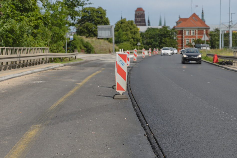 na zdjęciu jezdnia, po prawej widać nową nawierzchnię, na jezdni ustawione są też znaki drogowe, po prawej jadą samochody