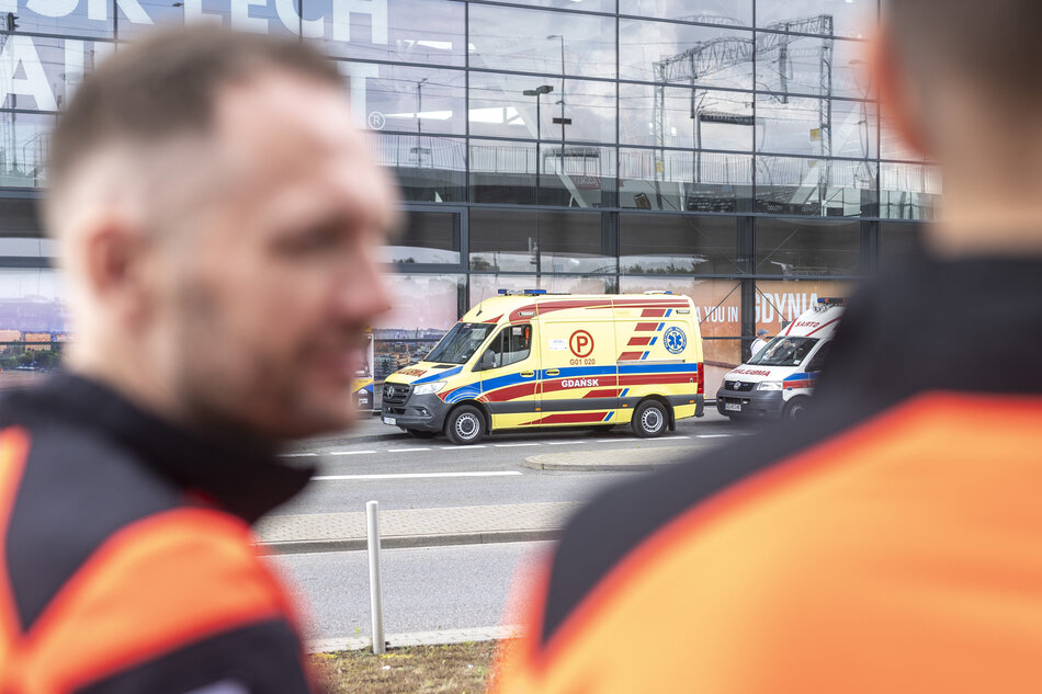 Na zdjęciu widać karetkę pogotowia ratunkowego z Gdańska stojącą przed nowoczesnym budynkiem ze szklaną fasadą. Na pierwszym planie rozmyte sylwetki dwóch ratowników medycznych w pomarańczowo-czarnych uniformach. 