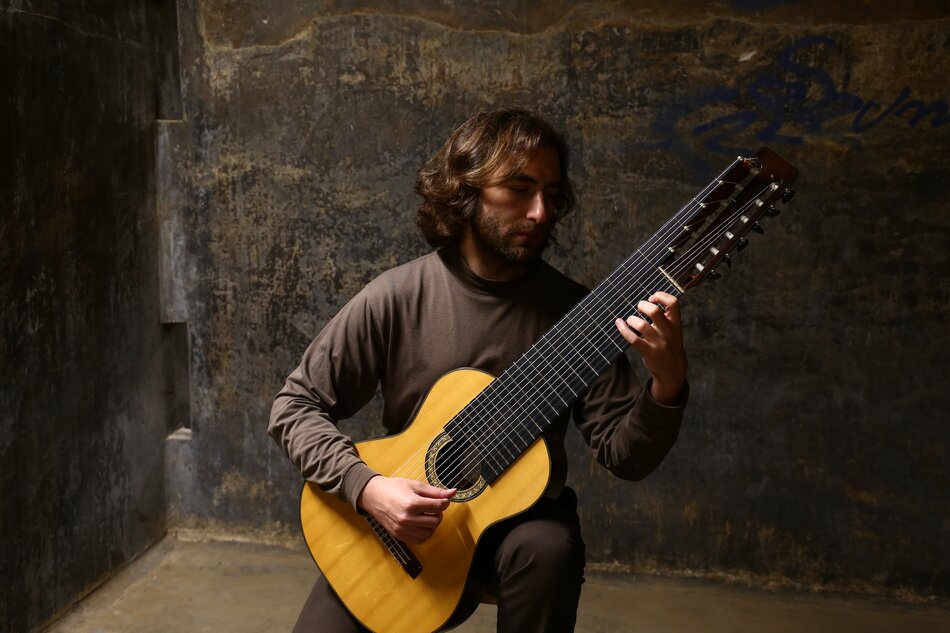 Na zdjęciu mężczyzna z włosami do ramion, z lekkim zarostem, trzymający w rękach stary instrument strunowy. 
