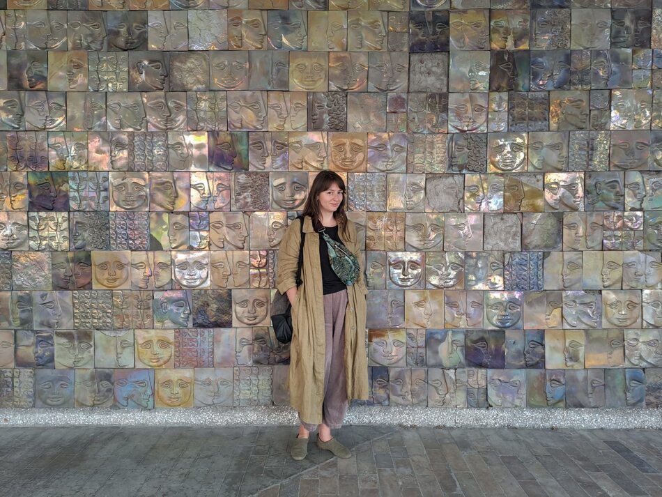 Na zdjęciu kobieta ubrana w jasnobrązowy płaszcz, stojąca na tle ściany z mozaiki