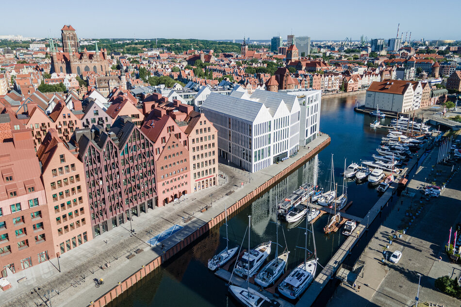 Zdjęcie ukazuje nabrzeże Wyspy Spichrzów w Gdańsku z nowoczesnymi apartamentowcami i mariną pełną jachtów. W tle rozciąga się panorama Głównego Miasta z wieżami kościołów i zabytkową zabudową. 