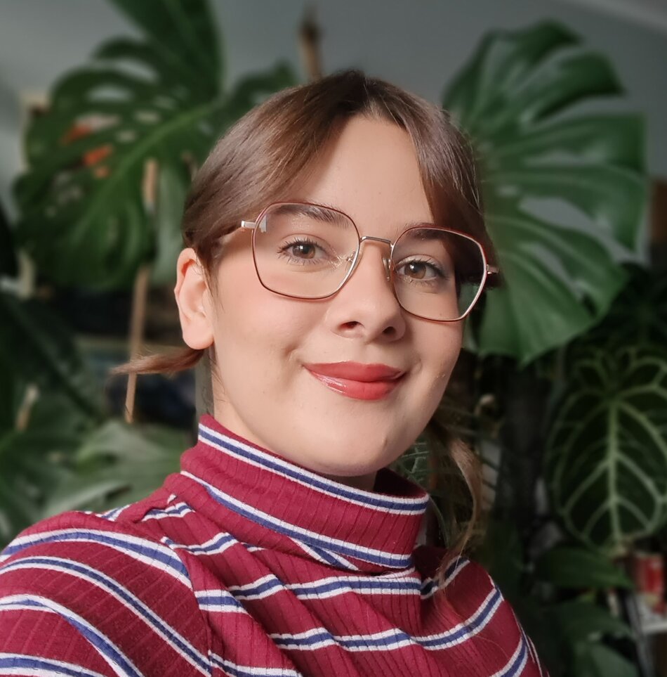 Na zdjęciu uśmiechnięta kobieta w okularach, w tle rośliny.