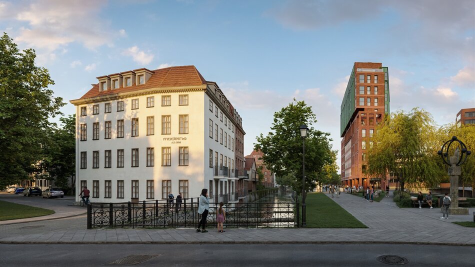 Na zdjęciu widzimy elegancką, spokojną przestrzeń miejską — najpewniej fragment Gdańska. Po lewej stronie znajduje się odnowiona kamienica w klasycznym stylu z białą elewacją i czerwonym dachem. Na fasadzie widnieje napis „Moderne A.D. 2024”, co wskazuje na nową inwestycję utrzymaną w historycznej stylistyce. Na prawo od niej biegnie kanał wodny, wzdłuż którego ciągną się brukowane alejki spacerowe. Przy kanale stoją ludzie — niektórzy spacerują, inni siedzą na ławkach, co tworzy sielską atmosferę. W tle, po prawej stronie, wyrasta nowoczesny budynek z czerwonej cegły z charakterystyczną zieloną nadbudową, łączący współczesną architekturę z gdańską tradycją.