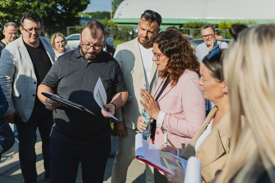 na zdjęciu grupa dorosłych osób stoi obok siebie, na pierwszym planie mężczyzna w średnim wieku w czarnej koszuli pokazuje coś na kartce kobiecie w różowej marynarce, także w średnim wieku
