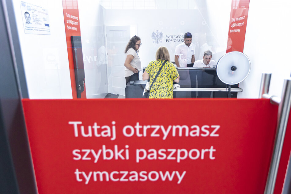 W punkcie obsługi paszportowej widoczna jest osoba składająca wniosek o szybki paszport tymczasowy, obsługiwana przez urzędników. Wnętrze punktu jest jasno oznaczone tablicami informacyjnymi i wyposażone w sprzęt do robienia zdjęć paszportowych. 