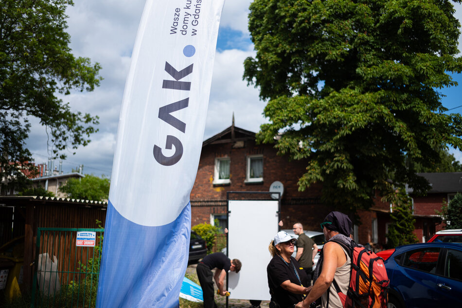 Na zdjęciu widać plenerową scenę z flagą GAK i kilkoma osobami rozmawiającymi na tle starej ceglanej zabudowy. Po lewej stronie widoczny jest baner instytucji, a w tle przygotowywana jest konstrukcja do wydarzenia na świeżym powietrzu.