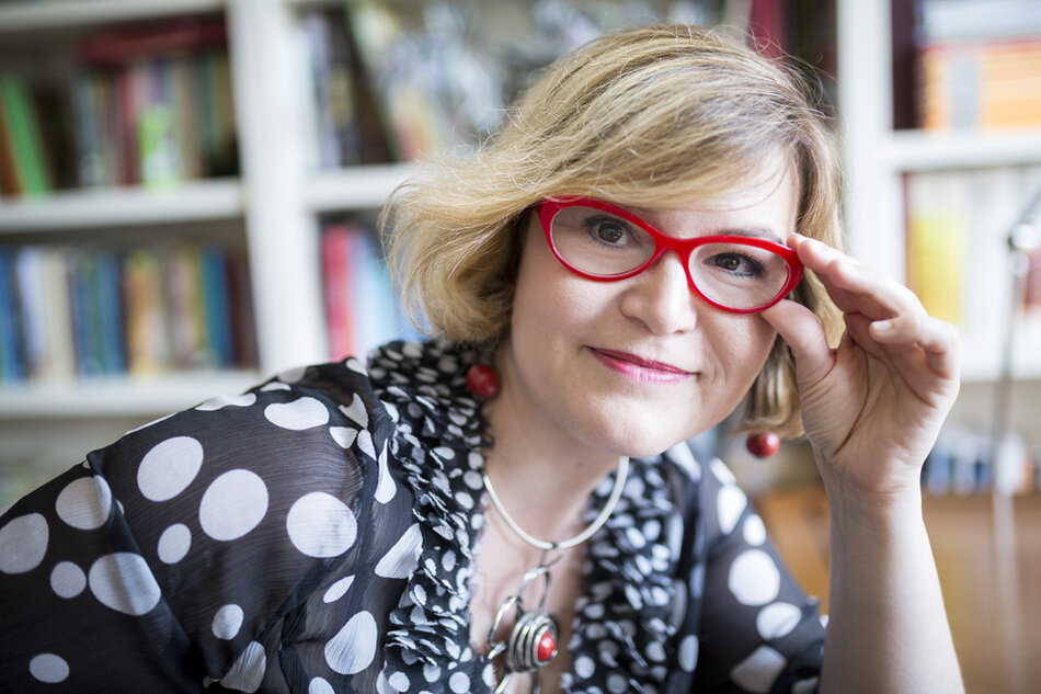 Na zdjęciu kobieta o krótkich blond włosach, w okularach z czerwonymi oprawkami, w tle regały z książkami.