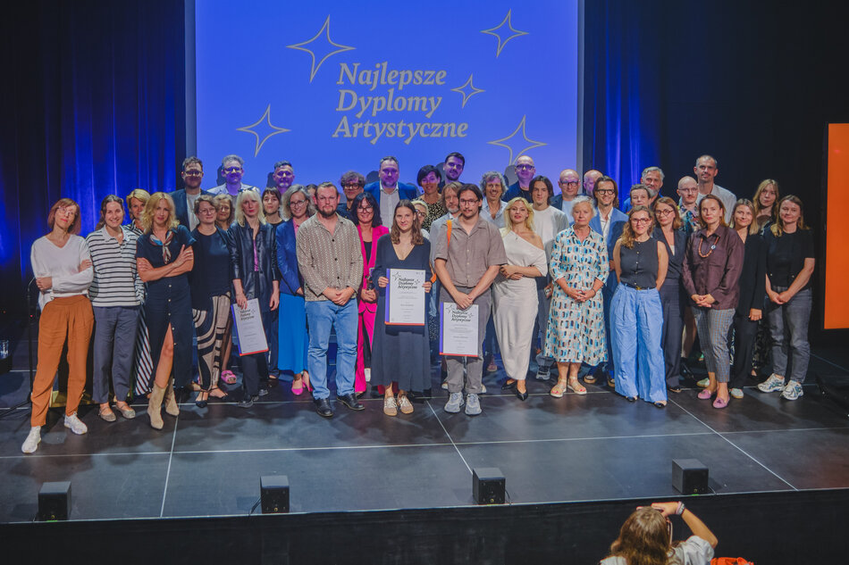 Na zdjęciu grupa osób stoi razem na scenie podczas uroczystości, na tle niebieskiej planszy z napisem „Najlepsze Dyplomy Artystyczne”. W centrum znajdują się laureaci trzymający dyplomy, otoczeni przez organizatorów i uczestników wydarzenia