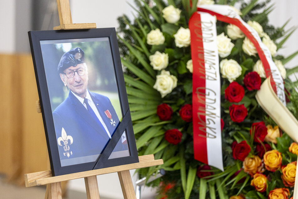 Na zdjęciu widoczna jest portretowa fotografia starszego mężczyzny w mundurze harcerskim z beretem i orderem, ustawiona na sztaludze i przewiązana czarną wstążką żałobną. Obok znajduje się wieniec z biało-czerwonymi kwiatami oraz szarfą z napisem Rada Miasta Gdańska, świadczący o uroczystości pogrzebowej. 