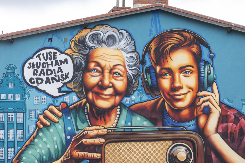 Zdjęcie ukazuje zbliżenie muralu, na którym starsza kobieta mówi „Tuse słucham Radia Gdańsk”, trzymając zabytkowe radio, a obok niej młody chłopak w słuchawkach uśmiecha się. Malowidło jest szczegółowe, wyraziste i celebruje 80-lecie Radia Gdańsk.