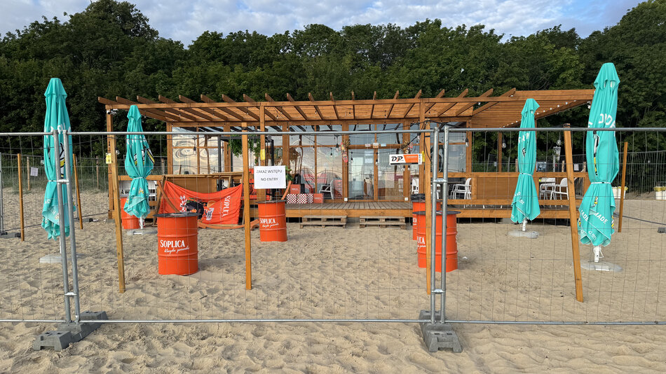 Na zdjęciu widoczna jest zamknięta strefa na plaży z drewnianym barem w tle, otoczona metalowym ogrodzeniem. Przed wejściem umieszczono tabliczkę z napisem „Zakaz wstępu”, a wokół stoją czerwone beczki z logo Soplica i zamknięte parasole turkusowe.