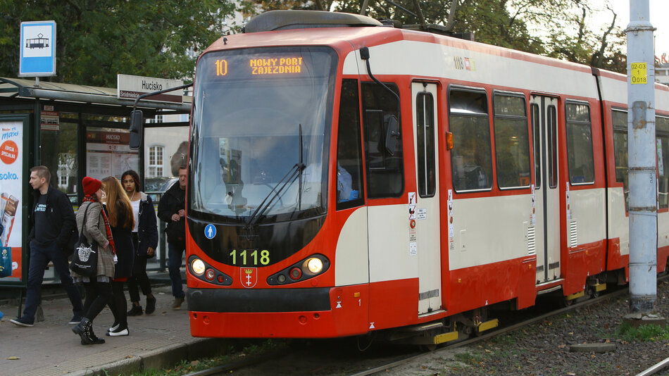 Tramwaj linii 10 zatrzymał się na przystanku Hucisko w Gdańsku, a pasażerowie wsiadają i wysiadają. Pojazd jest czerwono-biały, a w tle widać wiatę przystankową i tabliczkę z nazwą przystanku.