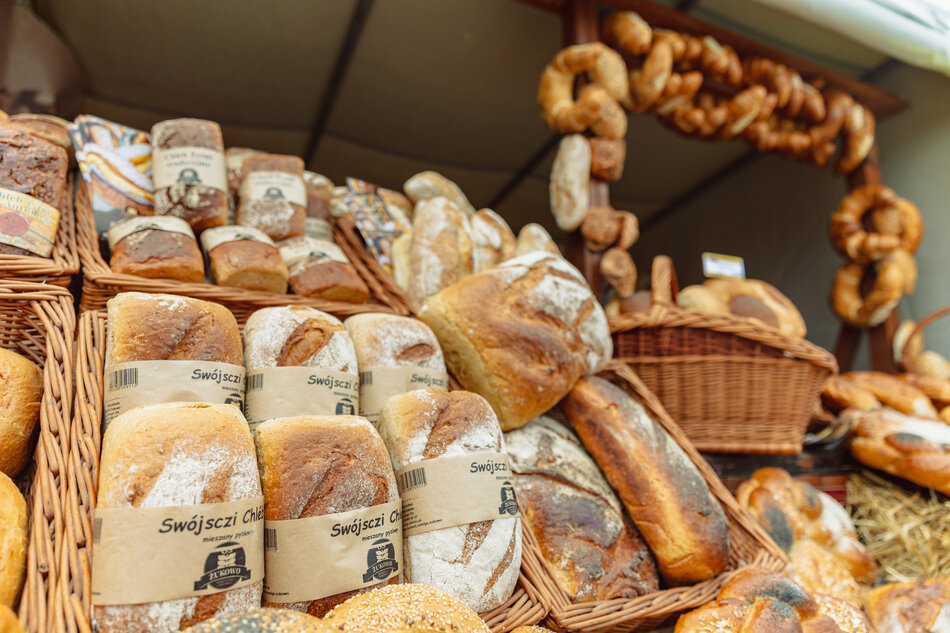 Zdjęcie przedstawia stoisko z tradycyjnym pieczywem na jarmarku lub festynie, pełne różnorodnych bochenków chleba i obwarzanków. Na pierwszym planie widoczne są chleby z etykietą „Swójszczi Chléb” z Żukowa, ułożone w wiklinowych koszach.