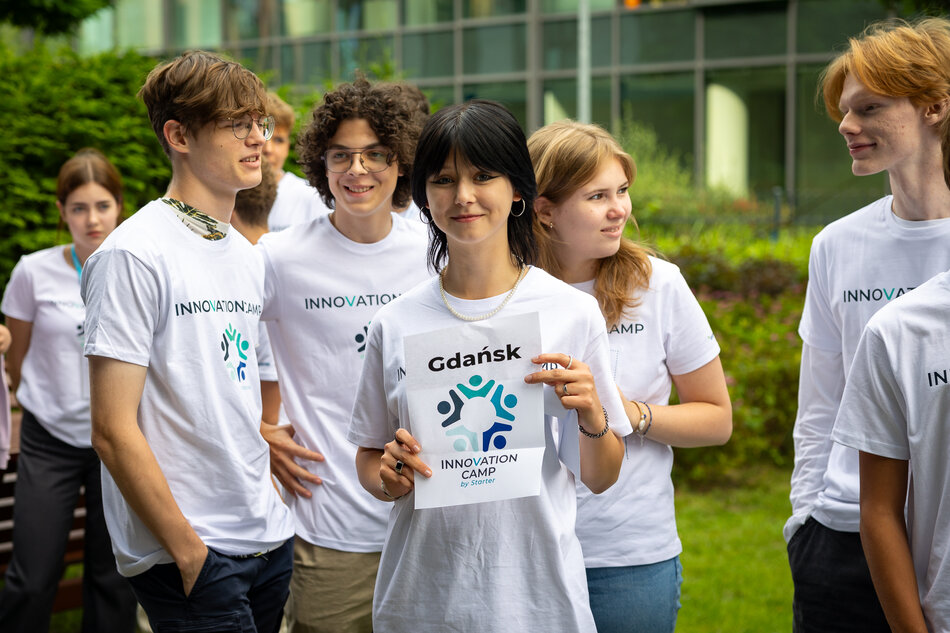 Grupa młodych osób w białych koszulkach z napisem „INNOVATION CAMP” pozuje razem na świeżym powietrzu. Dziewczyna na środku trzyma kartkę z napisem „Gdańsk” oraz logo wydarzenia, co sugeruje, że uczestniczą w innowacyjnym obozie organizowanym w tym mieście. 