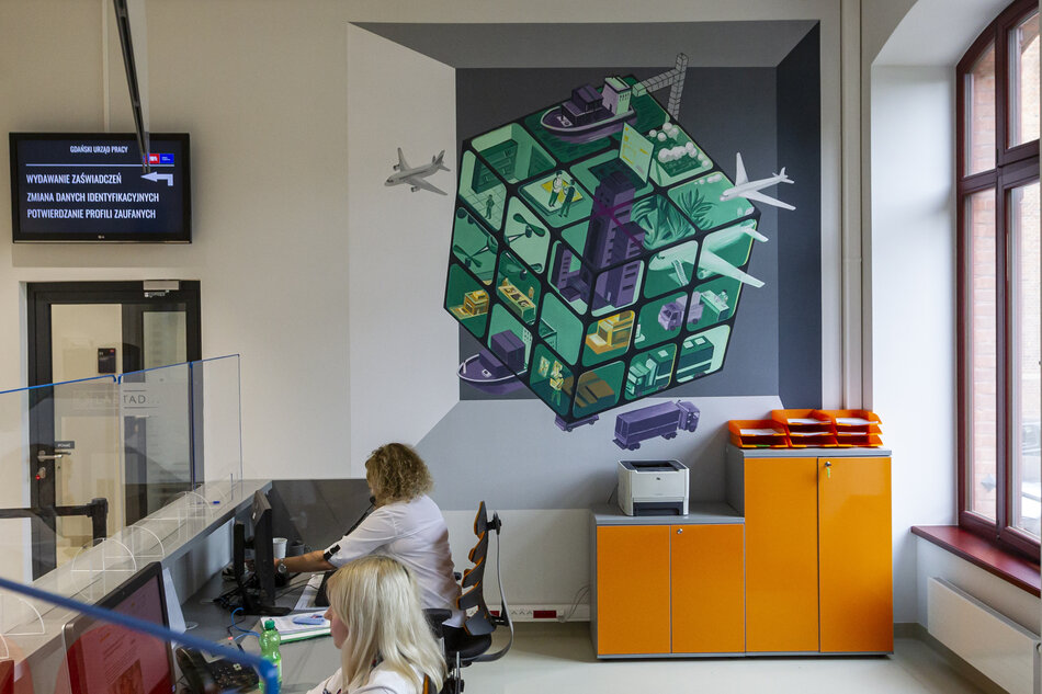 Zdjęcie przedstawia nowoczesne wnętrze urzędu, gdzie dwie pracownice obsługują petentów przy stanowiskach komputerowych. Na ścianie widoczny jest efektowny mural przedstawiający sześcienny labirynt z symbolami transportu, logistyki i przemysłu, nadający przestrzeni dynamiczny i nowoczesny charakter. 