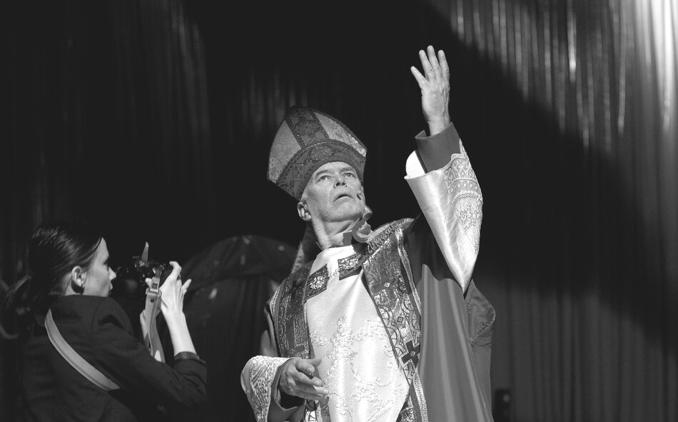 Na scenie widzimy aktora w roli duchownego, ubranego w bogato zdobione szaty liturgiczne i mitrę, unoszącego rękę w teatralnym geście. Z lewej strony kobieta z aparatem fotograficznym dokumentuje to dramatyczne ujęcie.