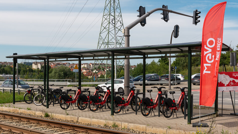 Na zdjęciu widoczna jest stacja rowerów miejskich MEVO zlokalizowana tuż przy torach kolejowych i sygnalizacji świetlnej, wyposażona w czerwono-czarne rowery elektryczne oraz kilka rowerów prywatnych. Obok stacji ustawiono czerwony baner z logo MEVO oraz tablicę informacyjną z mapą i listą nowych lokalizacji. 