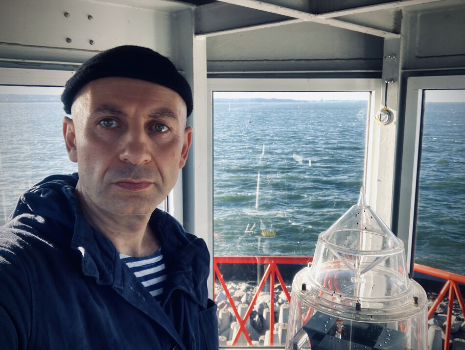 Na zdjęciu mężczyzna w czarnej czapeczce, znajduje się na mostku kapitańskim, za oknem fale morskie.