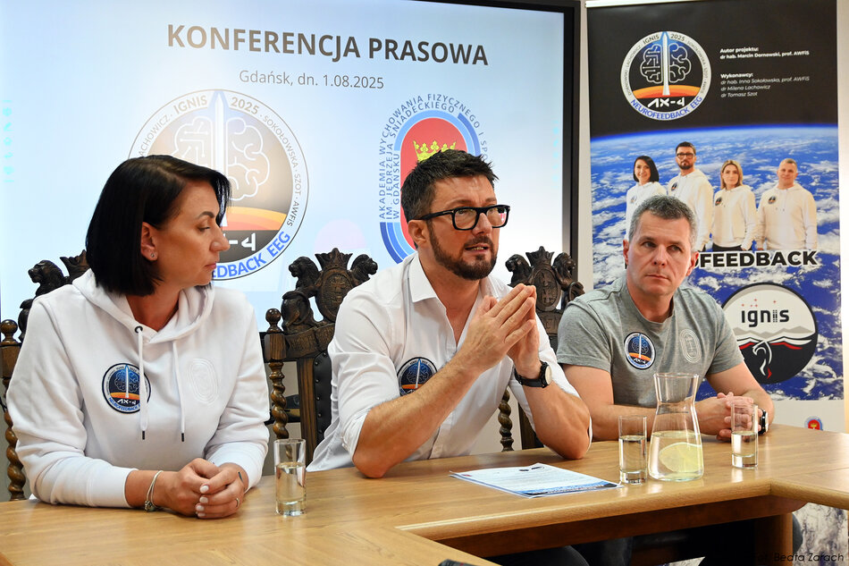 Jedna kobieta i dwóch mężczyzn siedzi za stołem podczas konferencji prasowej. Jeden mężczyzna mówi do mikrofonów.