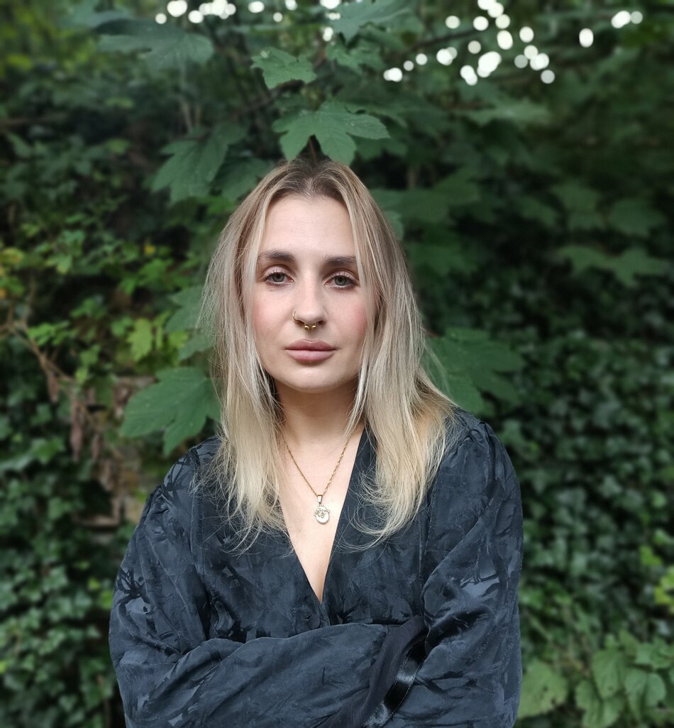 na zdjęciu kobieta z rozpuszczonymi włosami, za ramiona, na tle ściany liści.