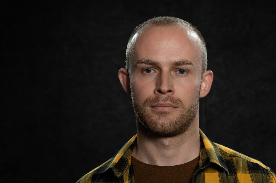 Na zdjęciu mężczyzna z bardzo krótkimi włosami, patrzący na wprost, w koszuli w kratę.