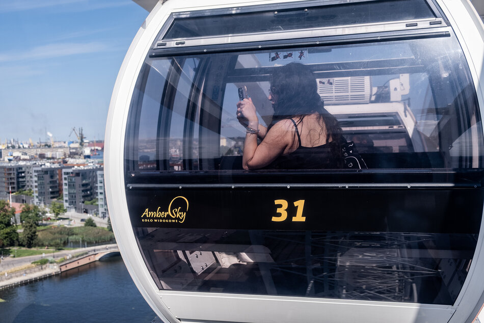 W gondoli koła widokowego tzw. diabelskiego młyna siedzi kobieta i robi zdjęcia przez okno telefonem. Za nią panorama miasta.