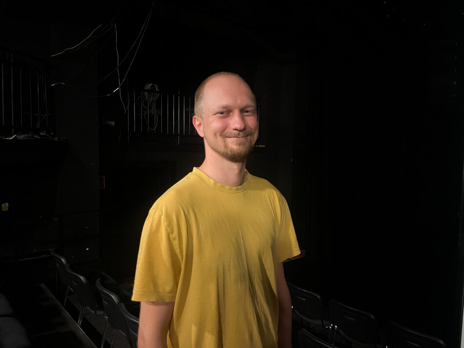 mężczyzna na czarnym tle, w żółtej koszulce, uśmiechnięty, z bardzo krótkimi włosami i lekkim zarostem