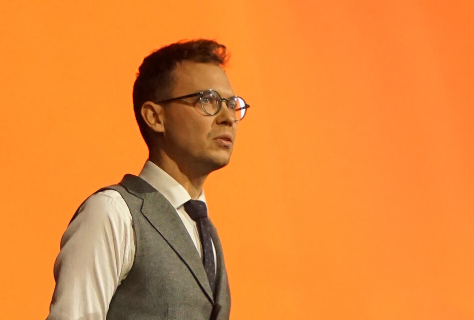 mężczyzna w koszuli z krawatem i kamizelce, z krótkimi włosami, w okularach, ustawiony bokiem na pomarańczowym tle 