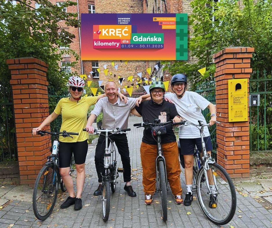 Cztery uśmiechnięte osoby stoją z rowerami przed wejściem do budynku z czerwonej cegły. Trzy z nich mają na głowach kaski rowerowe. Nad nimi zawieszony jest kolorowy baner z napisem ‘Kręć kilometry dla Gdańska, 01.09–30.11.2025’. W tle widać żółte chorągiewki rozwieszone na dziedzińcu.