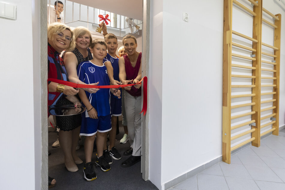 Na zdjęciu widać uroczyste przecięcie czerwonej wstęgi przez grupę osób, w tym dzieci w sportowych strojach oraz dorosłych, co symbolizuje otwarcie nowej przestrzeni. W tle ktoś trzyma ozdobny klucz z czerwoną kokardą, podkreślający wyjątkowy charakter wydarzenia. 