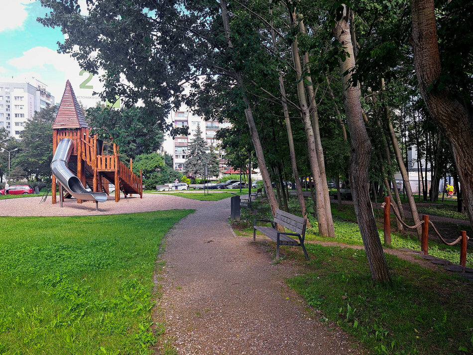 Na zdjęciu znajduje się park z alejką spacerową, ławkami i drzewami po prawej stronie. Po lewej widać duży, drewniany plac zabaw ze zjeżdżalnią, a w tle stoją bloki mieszkalne. 