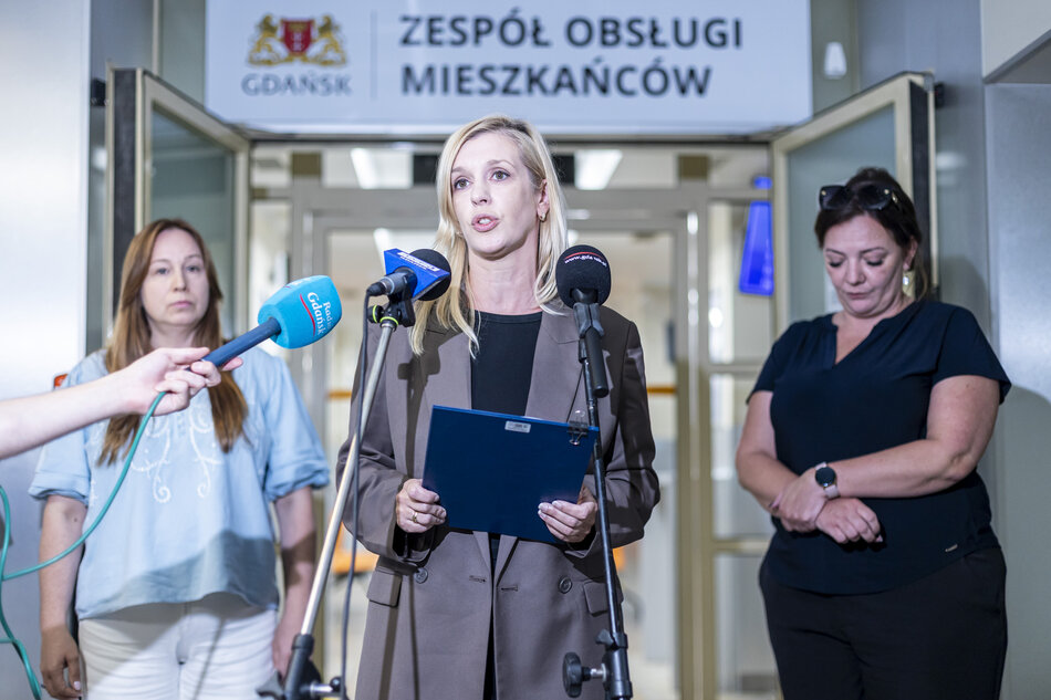 Na zdjęciu widać konferencję prasową przed wejściem do urzędu w Gdańsku, gdzie kobieta w marynarce przemawia do mediów, trzymając w ręku teczkę z dokumentami. Obok niej stoją dwie inne kobiety, a na mikrofonach widoczne są logotypy lokalnych stacji informacyjnych. 
