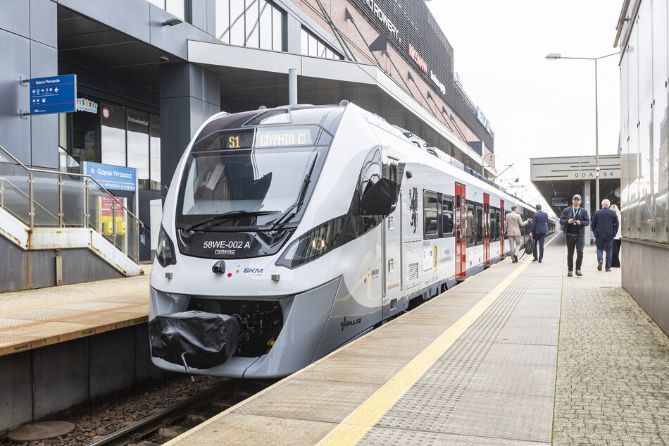 Na peronie stacji Gdańsk Wrzeszcz stoi nowoczesny pociąg SKM relacji S1 do Gdyni. Obok niego widać przechodniów oraz budynek galerii Metropolia z widocznymi szyldami sklepów. 