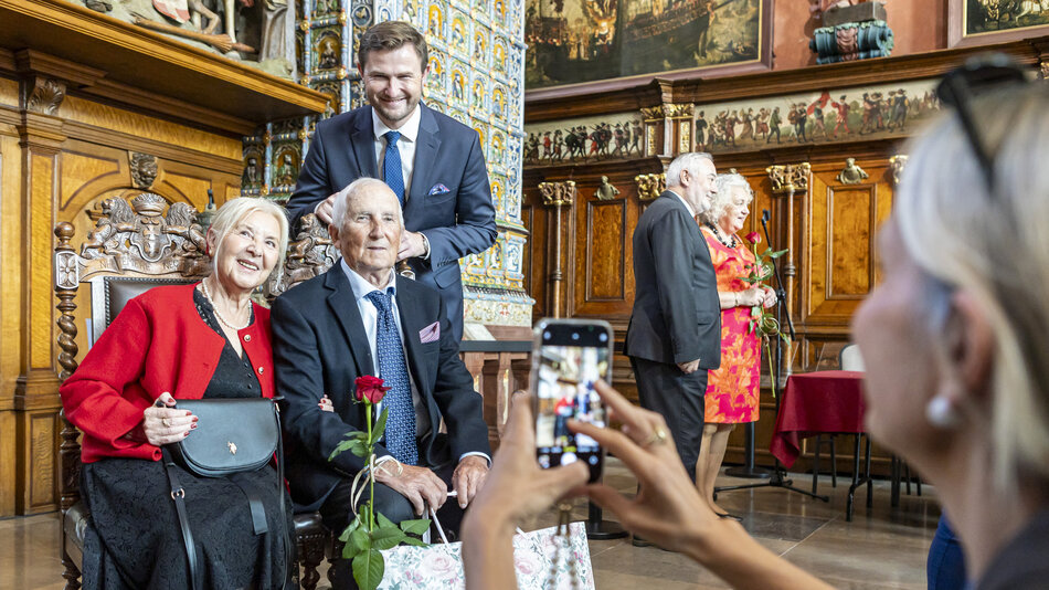grupa ludzi pozuje do zdjęcia, na tle pięknego zabytkowego wnętrza. W grupie ludzi na pierwszym planie siedzi para osób starszych, za nimi stoją trzy młodsze osoby w tym wysoki mężczyzna w garniturze. Na pierwszym planie widoczna kobieta robiąca zdjęcie telefonem