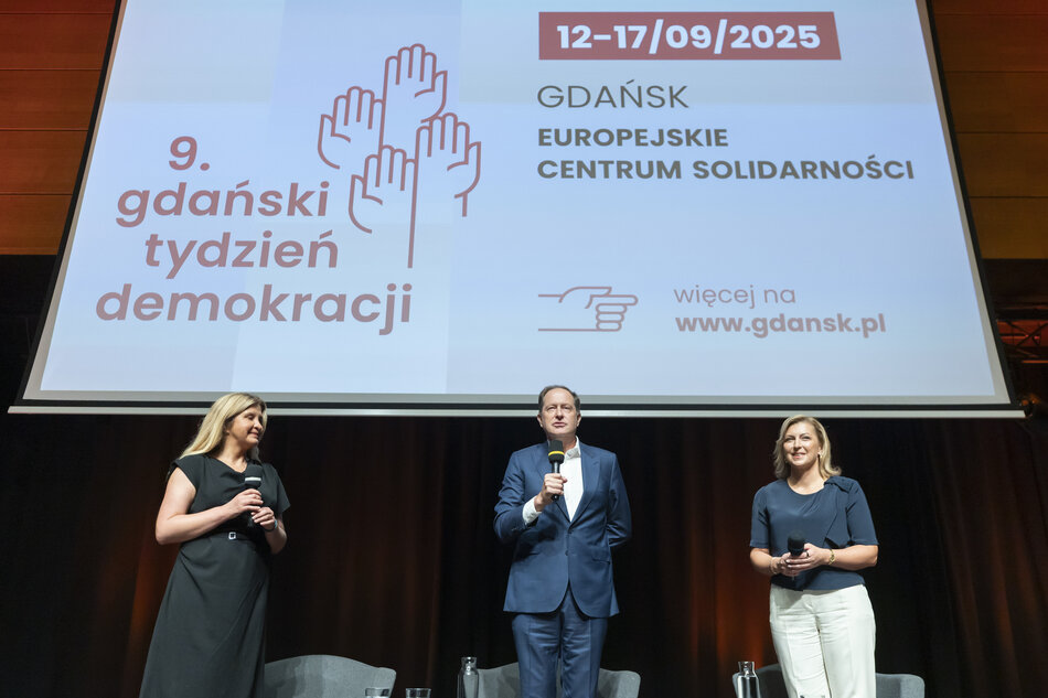  trzech osób stojących na scenie przed dużym ekranem. Na ekranie znajduje się tekst GDansk Europejskie Centrum Solidarności który tłumaczy się na 9. gdanskki tydzień demokracji w języku polskim. Poniżej ekranu znajdują się zdjęcia dwóch rąk trzymających mikrofon oraz data 12-17/09/2025.