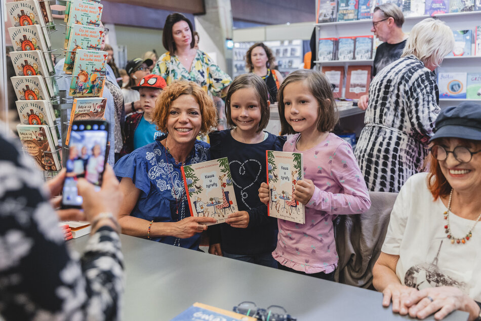 Na zdjęciu kobieta wraz z dwiema dziewczynkami pozuje do zdjęcia, trzymając książki pt. „Kawa u Żyrafy”. Scena odbywa się podczas targów książki, gdzie w tle widać stoisko z innymi publikacjami i odwiedzających. 