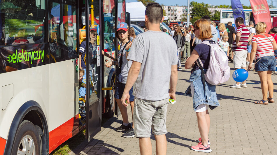 gcm-download-max-20230916-piknik-ztm-1-68c922a15bd5cgrupa ludzi stoi przed drzwiami małego autobusu. Na szybie pojazdu widoczny napis elektryczny. W tle widoczne kolorowe flagi i duże zgromadzenie ludzi