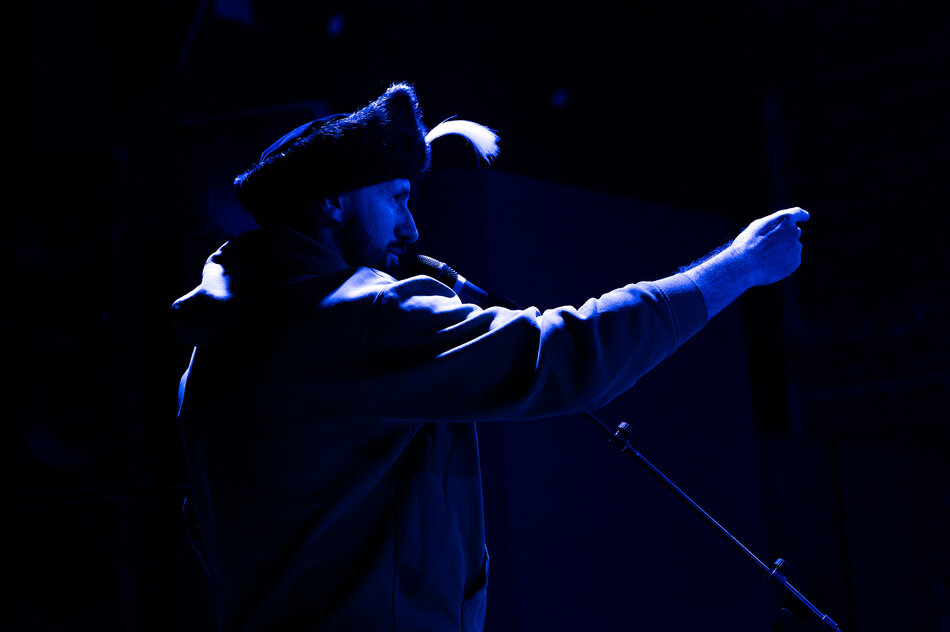 Na zdjęciu znajduje się osoba stojąca na scenie, oświetlona intensywnym niebieskim światłem. Ma na sobie bluzę z kapturem oraz czapkę z ozdobnym piórem. W jednej ręce trzyma mikrofon, a drugą gestykuluje, jakby przemawiała lub śpiewała do publiczności. Tło jest ciemne, niewyraźne, z widocznymi zarysami sprzętu scenicznego, takiego jak głośniki. Atmosfera zdjęcia jest dramatyczna i dynamiczna, sugerująca występ na żywo lub emocjonalne przemówienie.