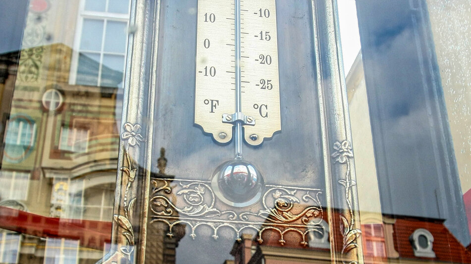 zdjęcie podwójnego termometru umieszczonego za szybą, w której odbijają się zabytkowe kamienice. Termometr ma dwie skale: Celsjusza i Fahrenheita