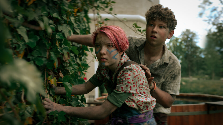 Dwójka młodych bohaterów z umalowanymi twarzami ukrywa się wśród liści bluszczu, wyglądając czujnie przed siebie. Ich ubrania są zabrudzone, co nadaje scenie atmosferę przygody lub zagrożenia.