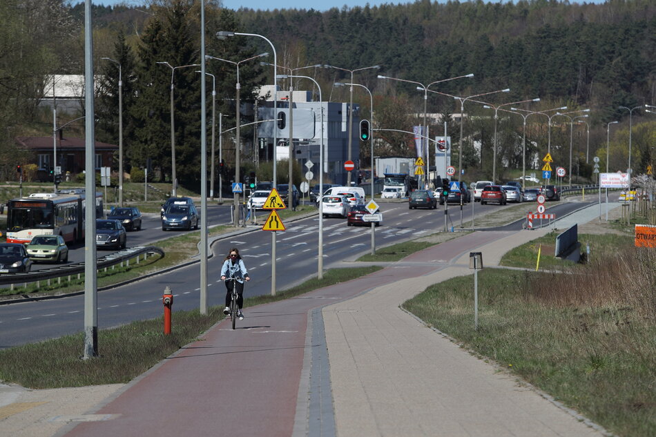 Na zdjęciu widzimy ruchliwą ulicę z kilkoma pasami ruchu, po której porusza się wiele samochodów i autobus. Na poboczu, równolegle do jezdni, biegnie ścieżka rowerowa, po której jedzie rowerzystka w jasnej kurtce. Obok ścieżki znajduje się chodnik dla pieszych. Widoczne są liczne znaki drogowe oraz sygnalizacja świetlna regulująca ruch. Po obu stronach drogi ustawione są latarnie. W tle widać zabudowania, a dalej rozciąga się las. Cała scena przedstawia fragment miasta z dobrze zorganizowaną infrastrukturą drogową, uwzględniającą zarówno samochody, jak i rowerzystów oraz pieszych.