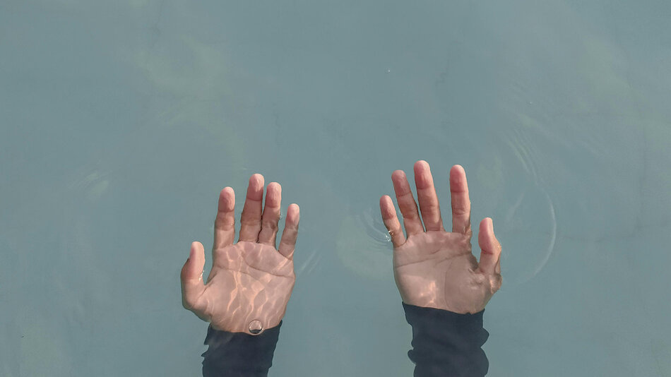 Dłonie zanurzone w wodzie unoszą się ku powierzchni, otoczone delikatnymi falami i odbiciami światła. Skóra widoczna jest wyraźnie pod przezroczystą taflą.