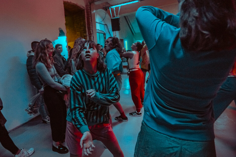 Na scenie performatywnej kilkanaście osób w jednakowych strojach przytula się i wspiera nawzajem, tworząc zwartą rzeźbiarską formę, podczas gdy inni widzowie siedzą przy ścianie obserwując spektakl.