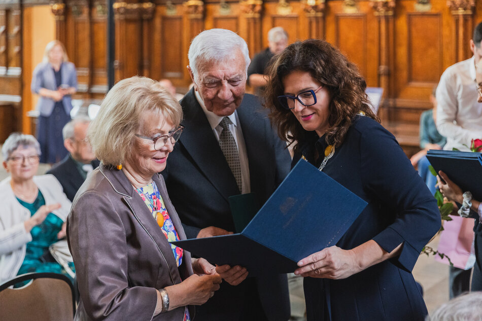 na zdjęciu dwoje seniorów małżeństwo pochyla się nad dyplomem który trzyma elegancko ubrana kobieta w średnim wieku