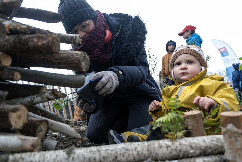 Malutkie dziecko w kombinezonie i czapeczce siedzi na ziemi, obok kuca młoda kobieta z sadzonką drzewa w rękach.