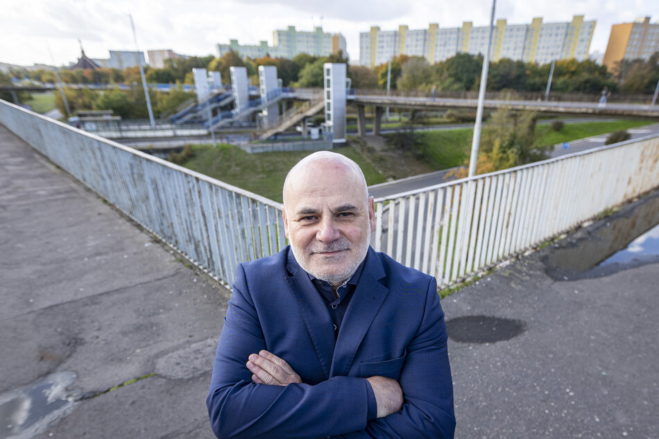 Mężczyzna w garniturze stoi na kładce dla pieszych z widokiem na osiedle mieszkaniowe i przystanek SKM w tle. Na jego twarzy widać pewność siebie, a tło ukazuje infrastrukturę miejską otoczoną jesienną zielenią. 