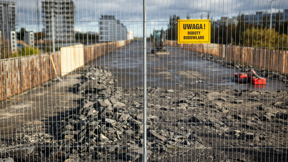 Zamknięty dla ruchu most jest w trakcie prac rozbiórkowych, o czym informuje żółta tabliczka z napisem „Uwaga! Roboty budowlane”. Na powierzchni widać stosy pokruszonego asfaltu i sprzęt budowlany w tle. 
