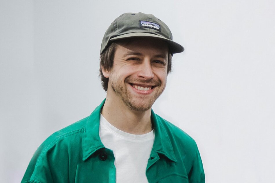zdjęcie przedstawia mężczyznę w czapce z daszkiem. Mężczyzna jest uśmiechnięty, ma na sobie zieloną koszulę oraz białą koszulkę.