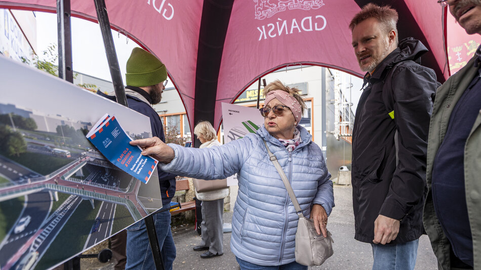 grupa ludzi stoi obok tablicy, na której widać wizualizację fragmentu ulicy i kładki pieszej. Nad nimi widać duży namiot i stojące obok flagi
